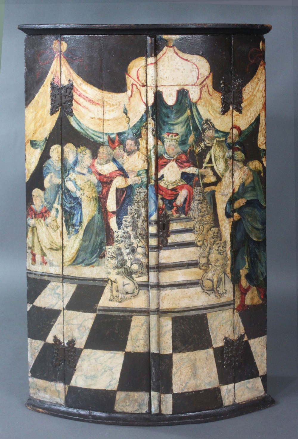An antique painted hanging corner cupboard featuring a detailed figurative scene with a crowned monarch, courtiers, drapery, and a chequerboard floor across its two doors.