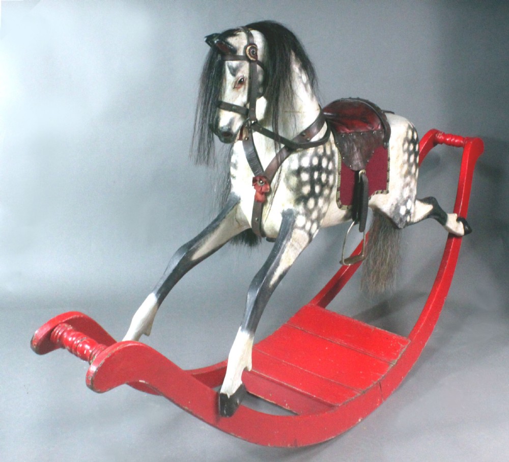 An antique carved wooden dapple grey rocking horse featuring a leather saddle, stirrups, black mane, and red curved bow rockers.