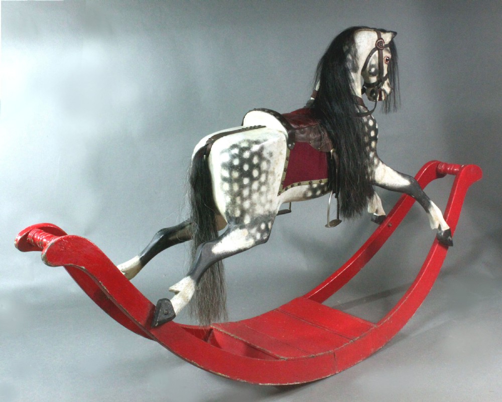 An antique carved wooden dapple grey rocking horse featuring a leather saddle, stirrups, black mane, and red curved bow rockers. View II