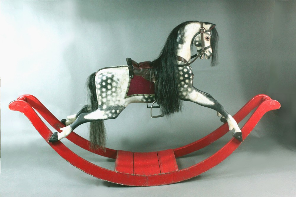 An antique carved wooden dapple grey rocking horse featuring a leather saddle, stirrups, black mane, and red curved bow rockers. View III