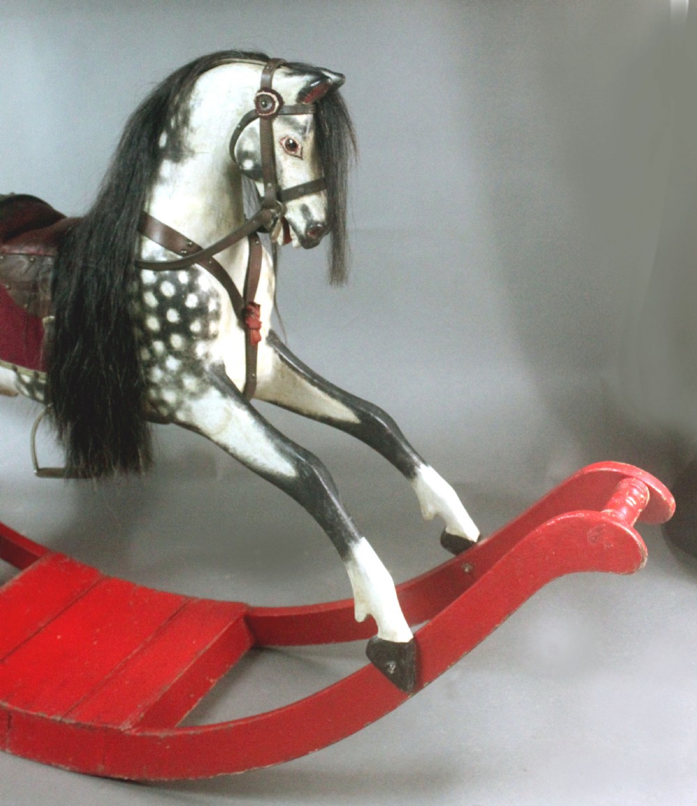 An antique carved wooden dapple grey rocking horse featuring a leather saddle, stirrups, black mane, and red curved bow rockers. View IV