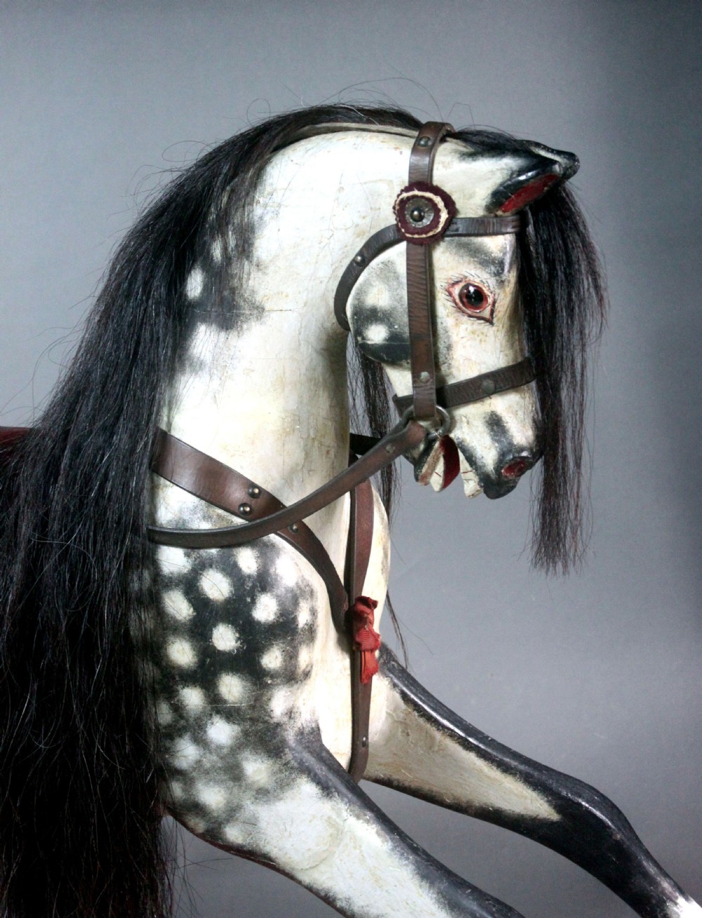 An antique carved wooden dapple grey rocking horse featuring a leather saddle, stirrups, black mane, and red curved bow rockers. View V