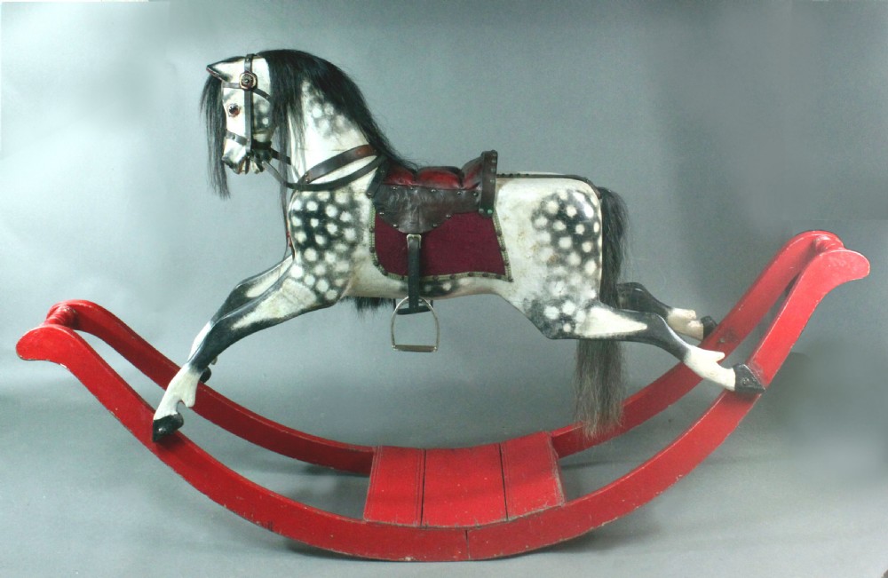 An antique carved wooden dapple grey rocking horse featuring a leather saddle, stirrups, black mane, and red curved bow rockers. View VI