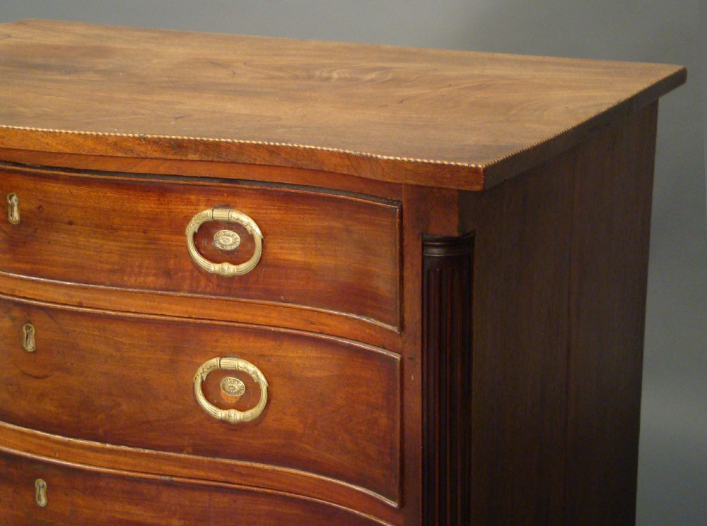 A dark brown mahogany Georgian chest of drawers featuring a shaped serpentine front, four graduated drawers with brass ring handles, and reeded corner columns on bracket feet. View II