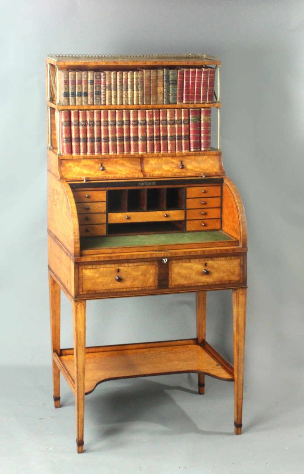 An antique satinwood cylinder bureau desk featuring an open two-tier bookshelf gallery at the top, a green leather writing surface inside, and tapered square legs with a low shelf.