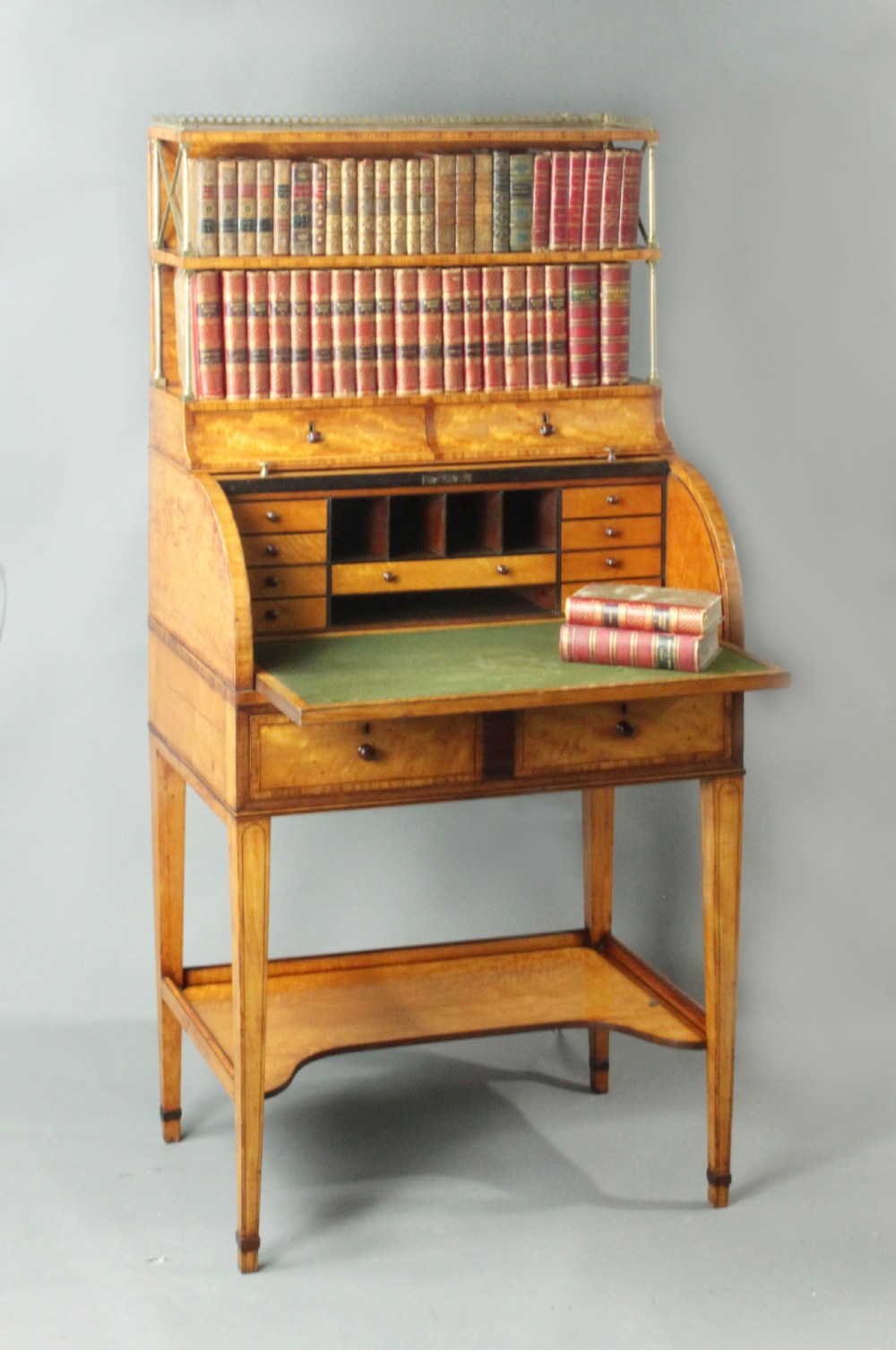 An antique satinwood cylinder bureau desk featuring an open two-tier bookshelf gallery at the top, a green leather writing surface inside, and tapered square legs with a low shelf. View II