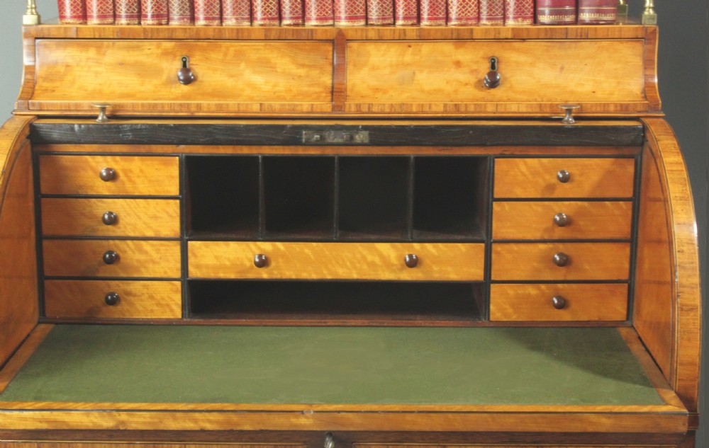 An antique satinwood cylinder bureau desk featuring an open two-tier bookshelf gallery at the top, a green leather writing surface inside, and tapered square legs with a low shelf. View IV