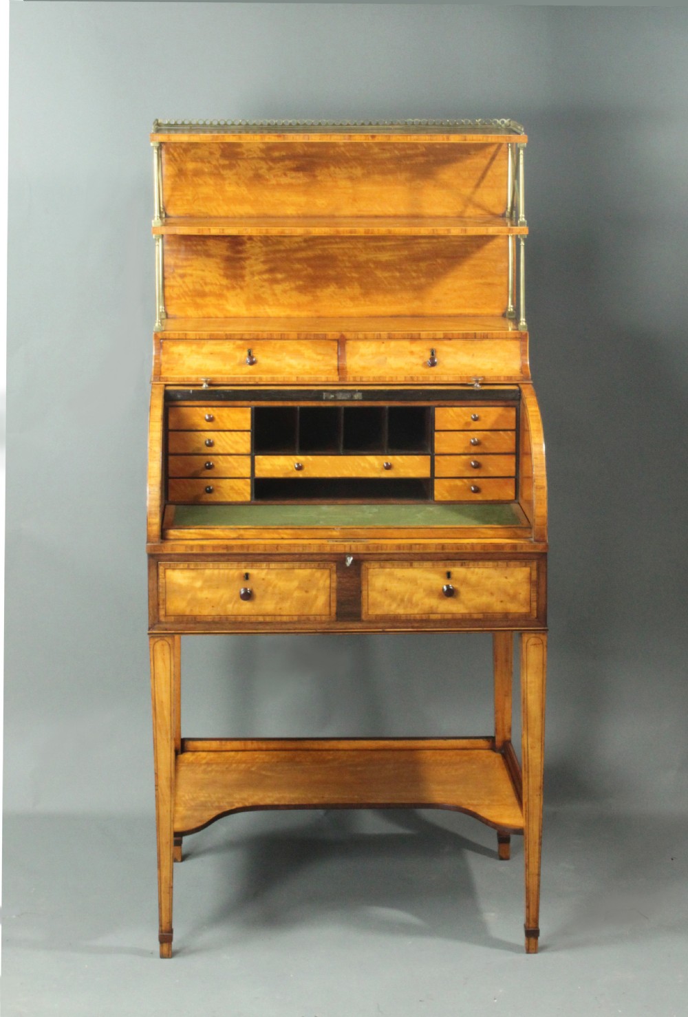 An antique satinwood cylinder bureau desk featuring an open two-tier bookshelf gallery at the top, a green leather writing surface inside, and tapered square legs with a low shelf. View V