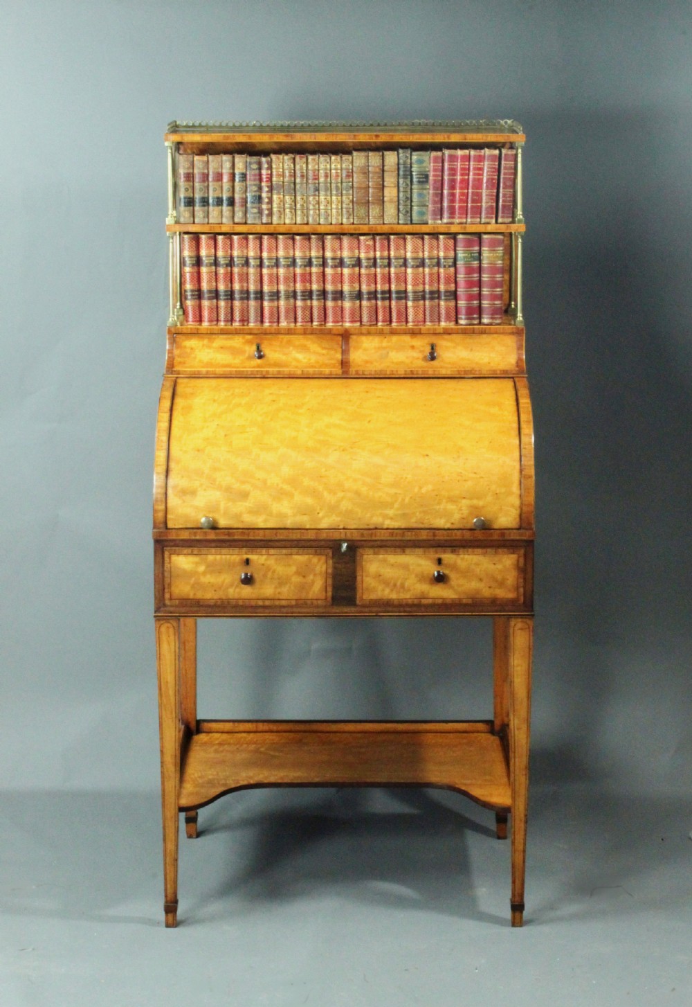 An antique satinwood cylinder bureau desk featuring an open two-tier bookshelf gallery at the top, a green leather writing surface inside, and tapered square legs with a low shelf. View VI