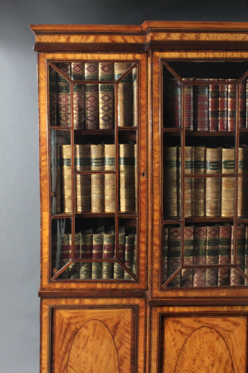 A four-door Georgian breakfront bookcase crafted from golden satinwood featuring geometric astragal glazing bars above four oval inlaid cupboard doors. View VII