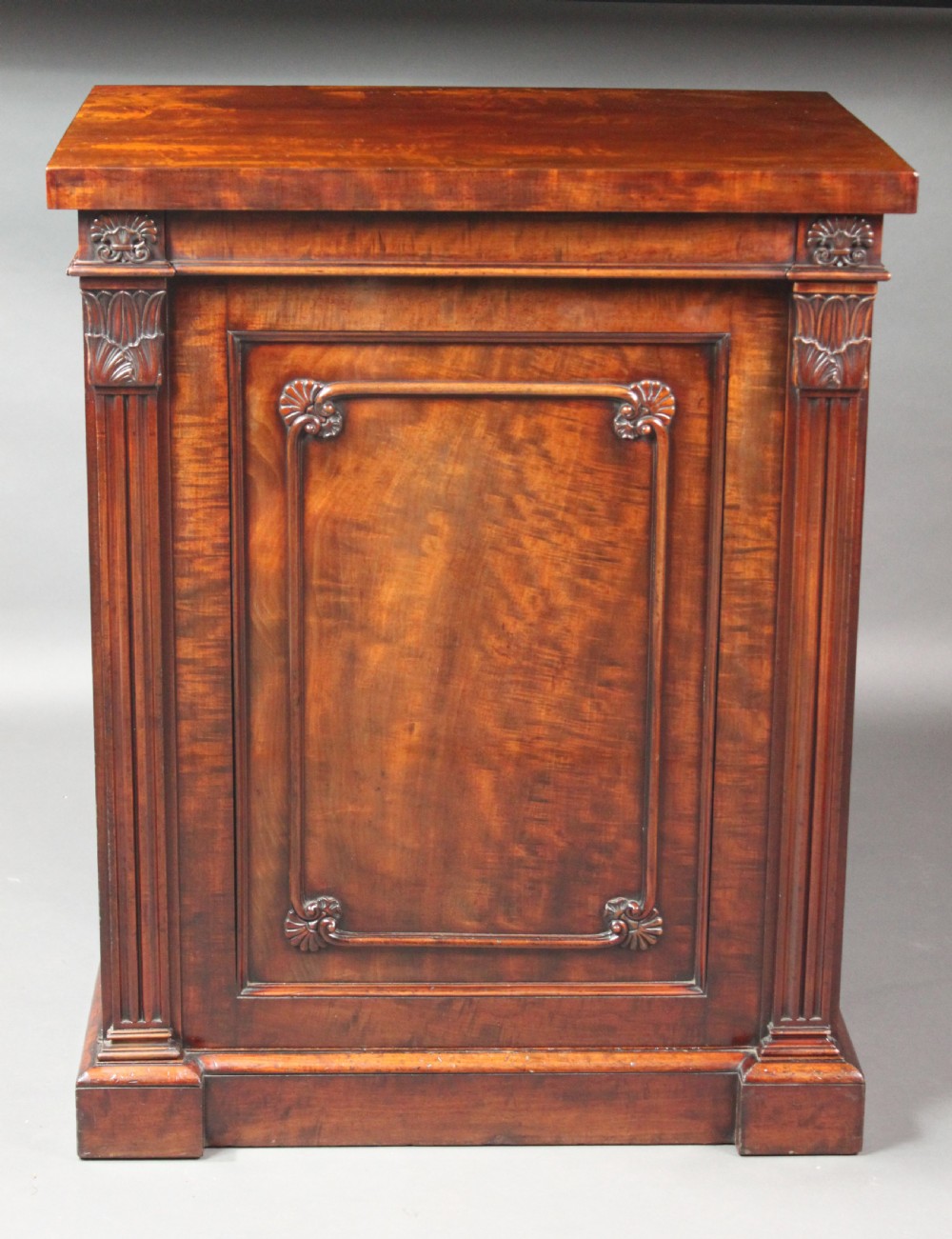 A nineteenth-century flame mahogany library cabinet featuring carved pilasters, reeded side columns, and a single recessed panel door set on a solid plinth base. View V