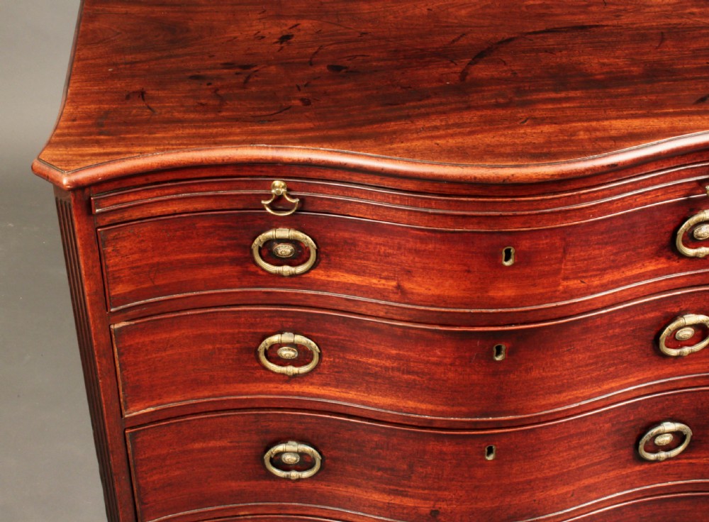 An antique George III mahogany chest of drawers featuring a curved serpentine front, four graduated drawers with brass handles, and sturdy bracket feet. View III