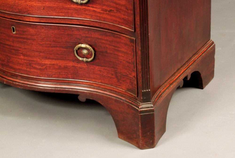 An antique George III mahogany chest of drawers featuring a curved serpentine front, four graduated drawers with brass handles, and sturdy bracket feet. View IV