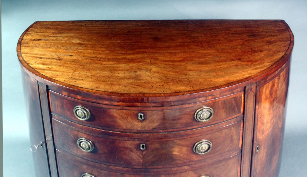 A late 18th-century George III flame mahogany demi-lune commode featuring four central drawers flanked by side cupboards, resting on bracket feet. View XII