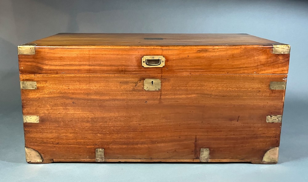 An antique rectangular camphor wood campaign chest featuring recessed brass handles, corner brackets, and a central brass escutcheon.