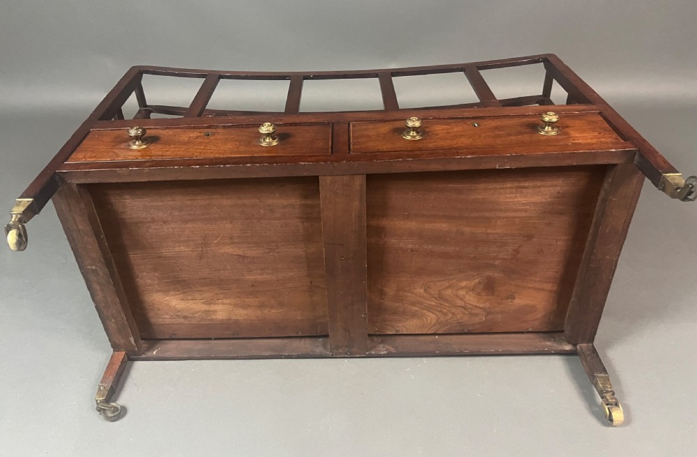 An antique Georgian mahogany Canterbury featuring a slatted concave top section, four internal compartments, two lower drawers with brass knobs, and slim tapered legs on brass castors. View IX