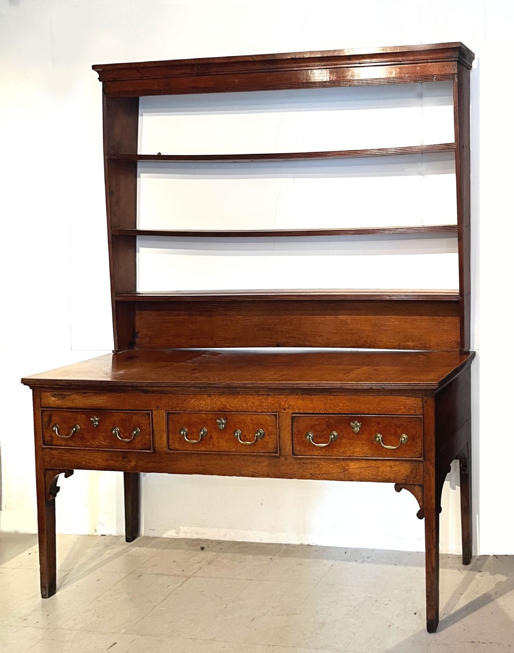 An antique oak dresser featuring a three-drawer base with brass swan-neck handles, topped with a tall open three-shelf plate rack.