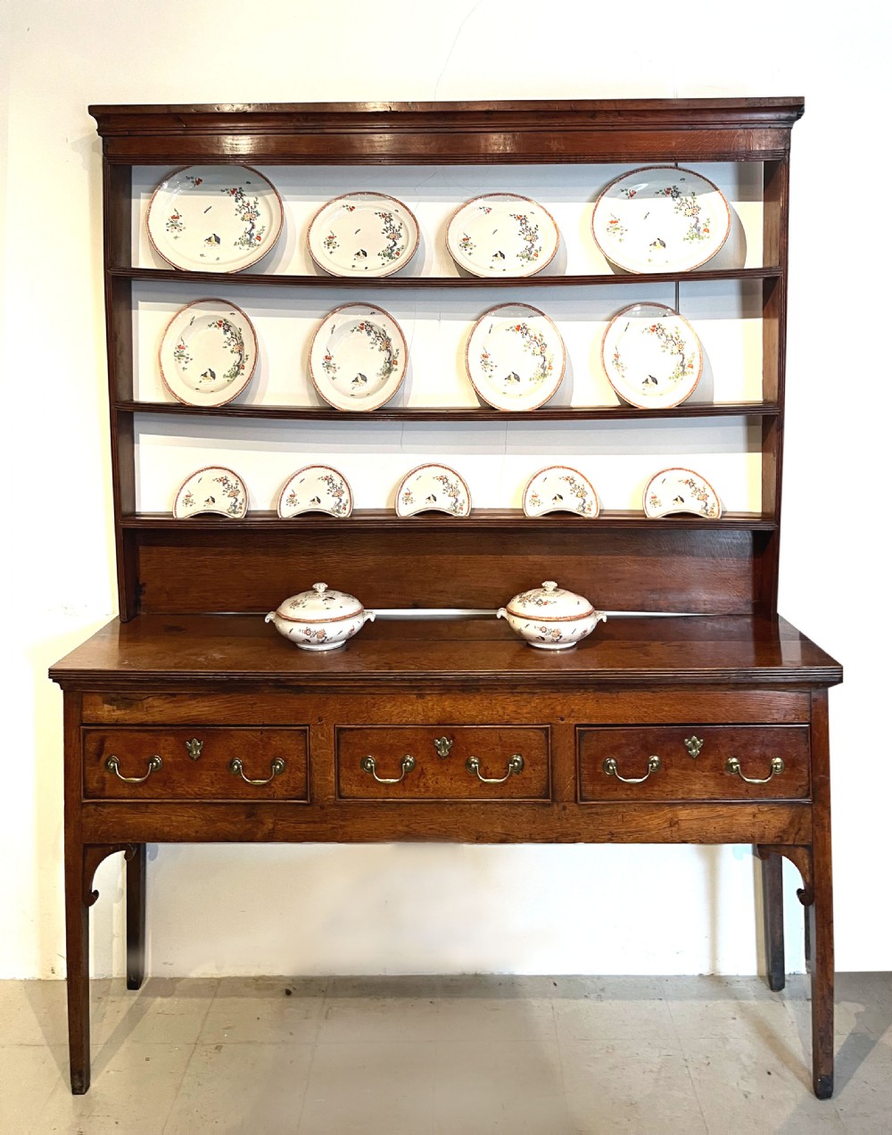 An antique oak dresser featuring a three-drawer base with brass swan-neck handles, topped with a tall open three-shelf plate rack. View II