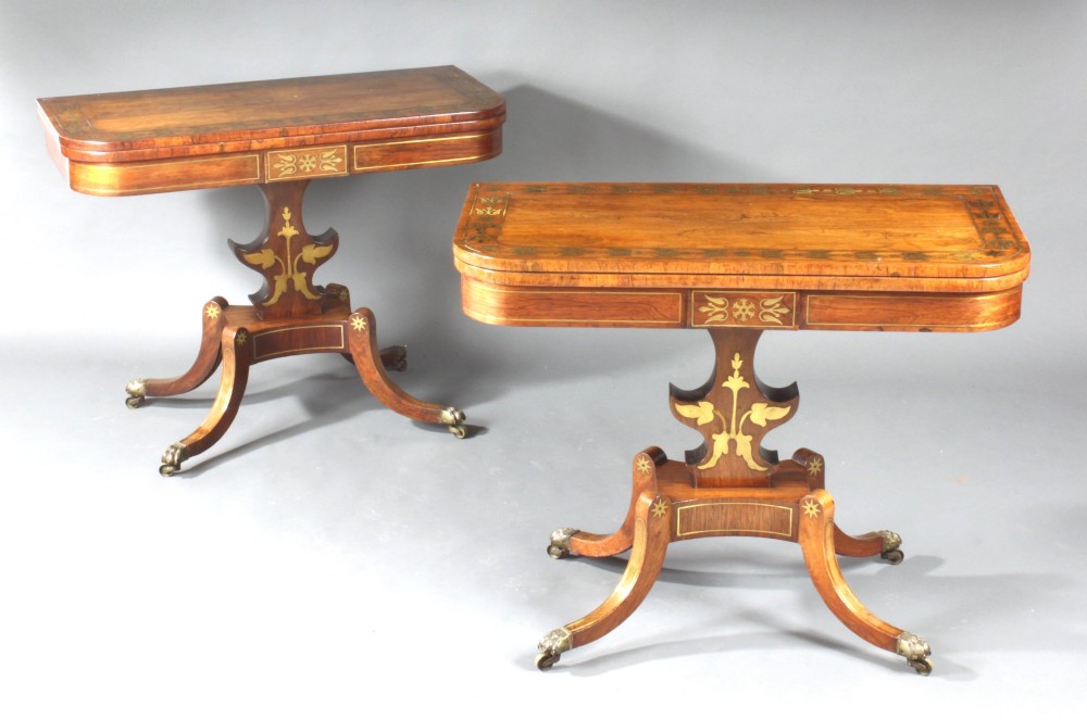 A pair of Regency rosewood card tables featuring brass foliate inlay, fold-over tops, pedestal bases, and splayed legs with brass paw castors.