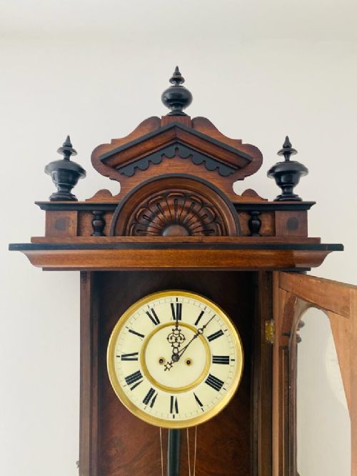 fig 1. A dark brown glazed wooden Vienna regulator wall clock featuring carved cresting, turned side columns, a white Roman numeral dial, and brass weights with a pendulum.