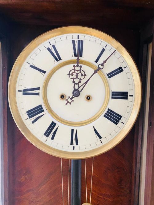 fig 2. A dark brown glazed wooden Vienna regulator wall clock featuring carved cresting, turned side columns, a white Roman numeral dial, and brass weights with a pendulum.