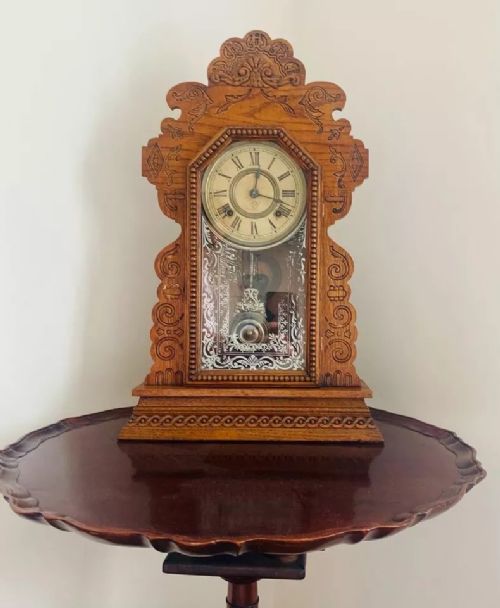fig 1. An antique American Ansonia gingerbread mantel clock featuring a pressed oak case with scrollwork crest, Roman numeral dial, and stencilled glass door revealing the brass pendulum.