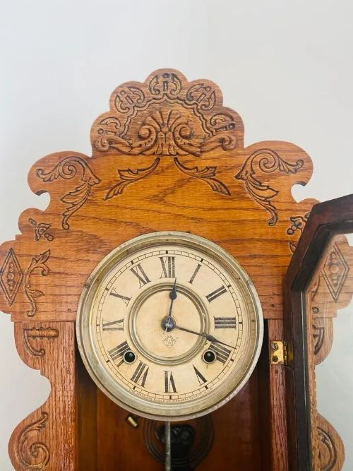 fig 2. An antique American Ansonia gingerbread mantel clock featuring a pressed oak case with scrollwork crest, Roman numeral dial, and stencilled glass door revealing the brass pendulum.