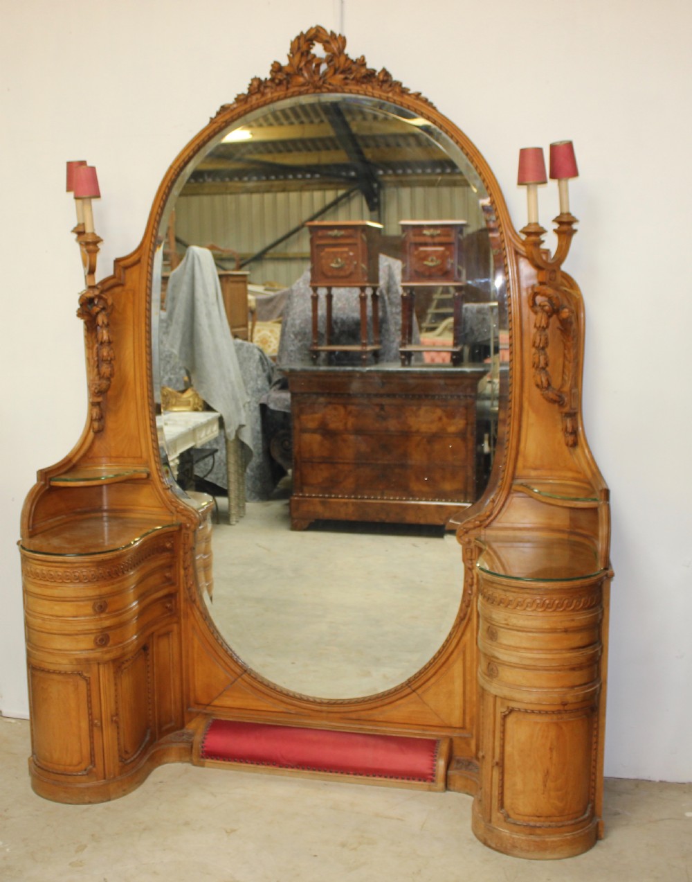 A French carved walnut dressing table featuring a large central oval mirror, twin side sconces with fabric shades, pedestals with glass tops, and a low red velvet footrest.