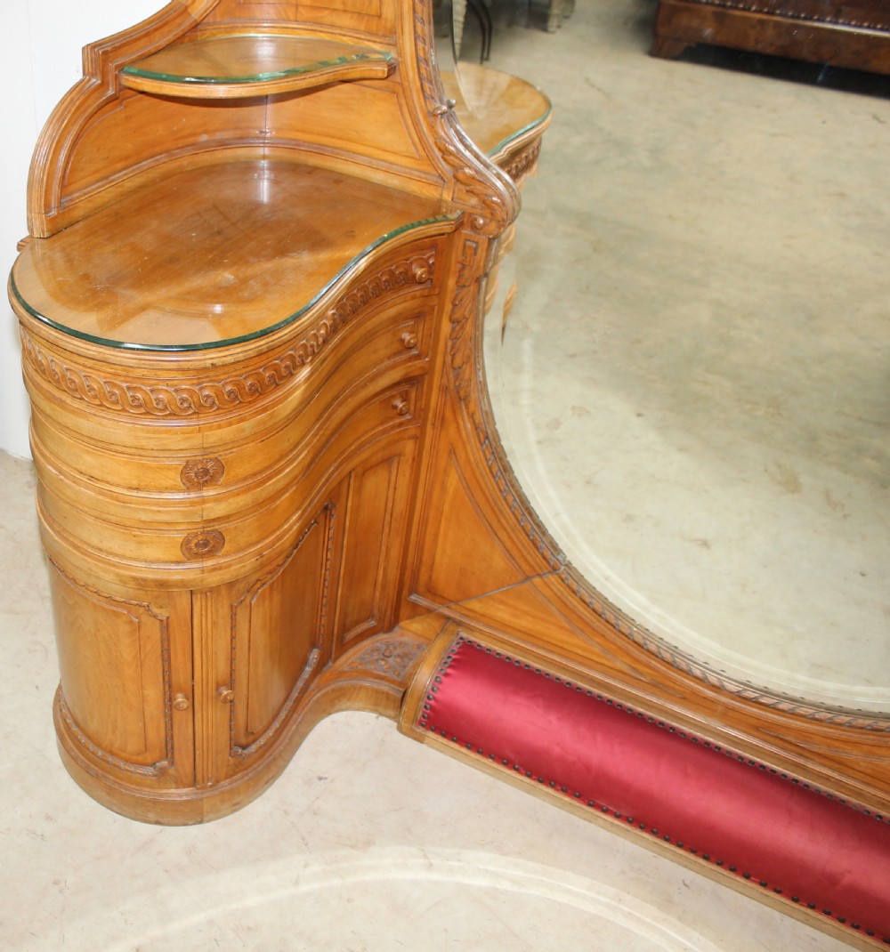 A French carved walnut dressing table featuring a large central oval mirror, twin side sconces with fabric shades, pedestals with glass tops, and a low red velvet footrest. View V
