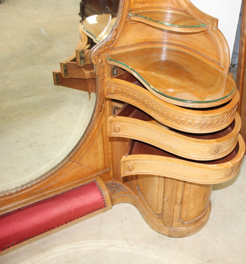 A French carved walnut dressing table featuring a large central oval mirror, twin side sconces with fabric shades, pedestals with glass tops, and a low red velvet footrest. View VIII