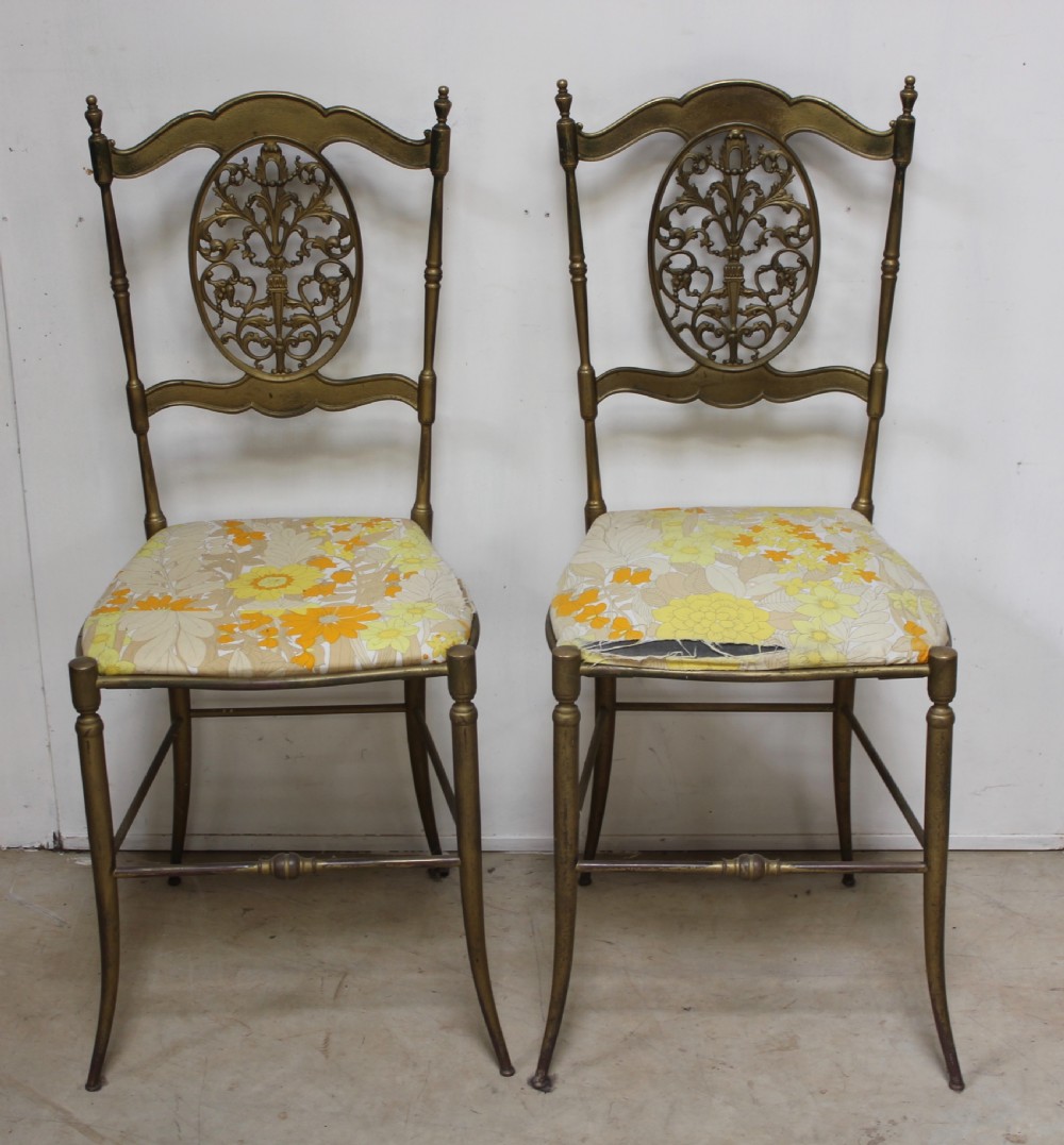 A pair of gold-toned brass side chairs featuring oval openwork splat backs, turned stretchers, and yellow floral fabric seats.