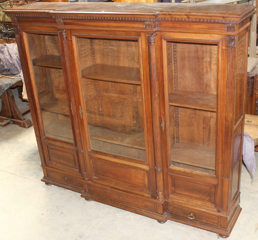 A large antique warm brown wooden breakfront bookcase with three glazed doors, carved fluted pilasters, and lower drawers.