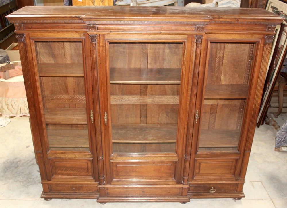 A large antique warm brown wooden breakfront bookcase with three glazed doors, carved fluted pilasters, and lower drawers. View II