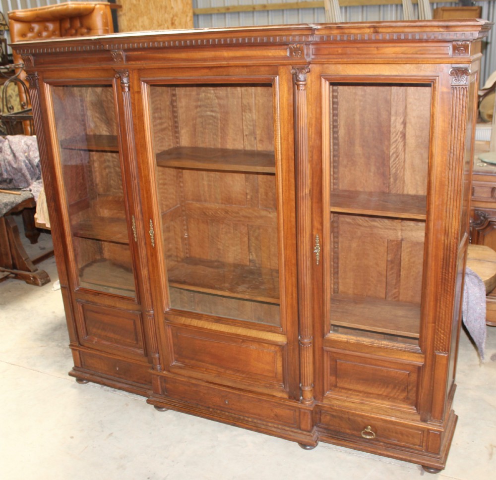 A large antique warm brown wooden breakfront bookcase with three glazed doors, carved fluted pilasters, and lower drawers. View III