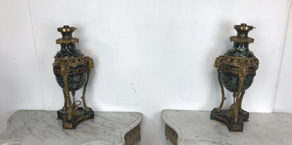 A pair of Neoclassical style table lamps fashioned as dark green marble urns supported by gilt bronze triform legs featuring rams head mounts.