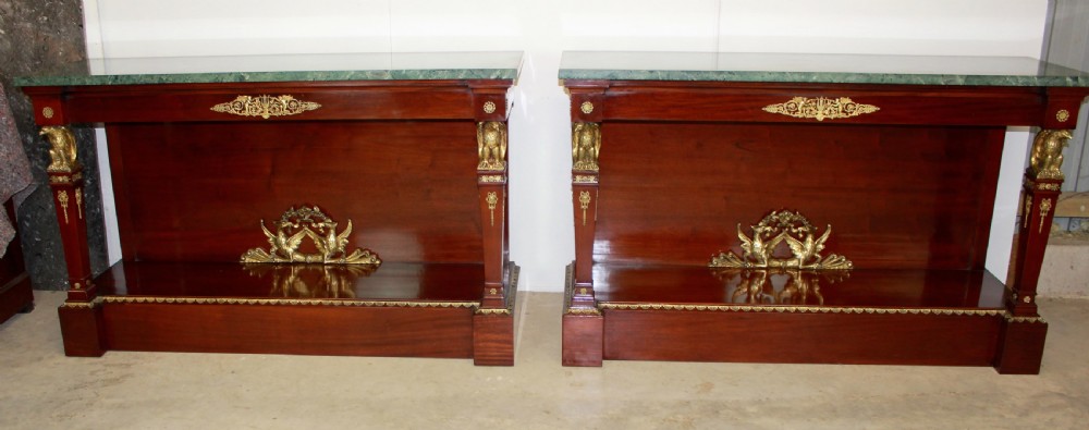 A pair of dark mahogany French Empire console tables featuring green marble tops, winged eagle supports, lower plinth shelves, and ornate gilt bronze mounts.