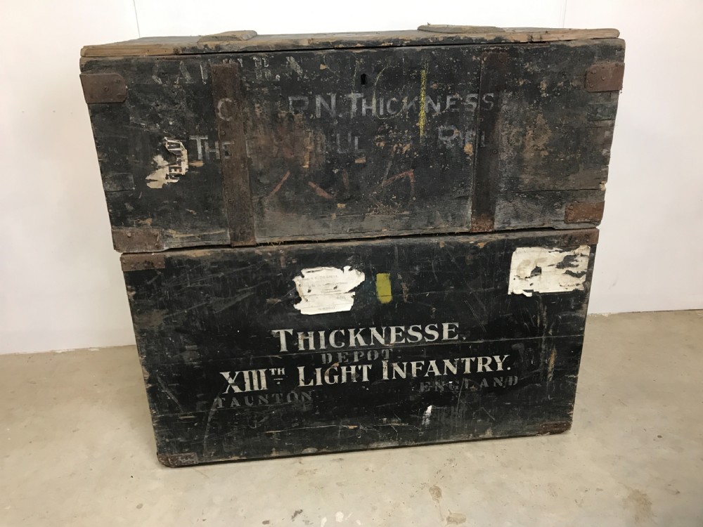 Two stacked dark painted antique wooden military trunks featuring original stencilled regimental lettering and iron brackets.
