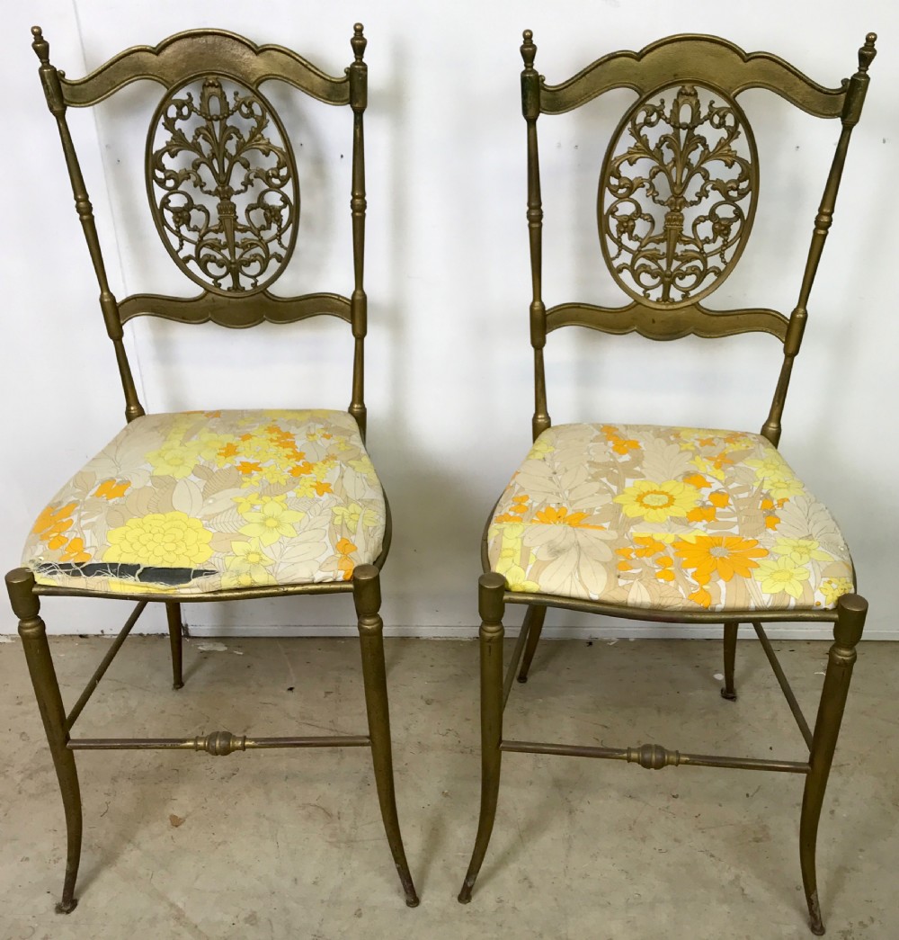 A pair of solid brass side chairs featuring ornate openwork oval backrests, slender turned legs, and yellow floral upholstered seats.