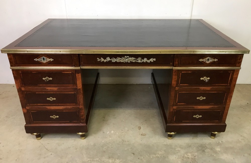 An antique mahogany pedestal desk featuring a dark leather writing top, multiple drawers with brass keyhole escutcheons, and central metal mounts.