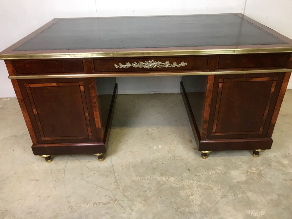 An antique mahogany pedestal desk featuring a dark leather writing top, multiple drawers with brass keyhole escutcheons, and central metal mounts. View X