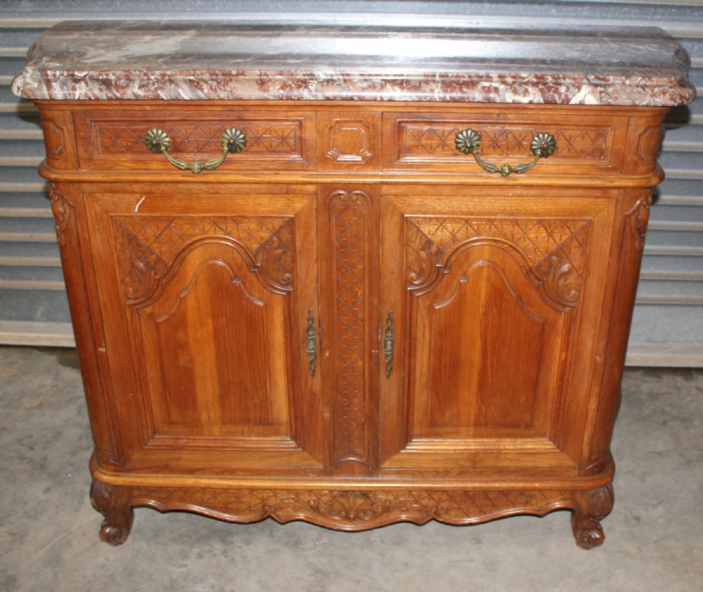 A carved French provincial oak sideboard featuring two drawers, two cabinet doors, carved geometric and floral motifs, cabriole feet, and a thick grey veined marble top.