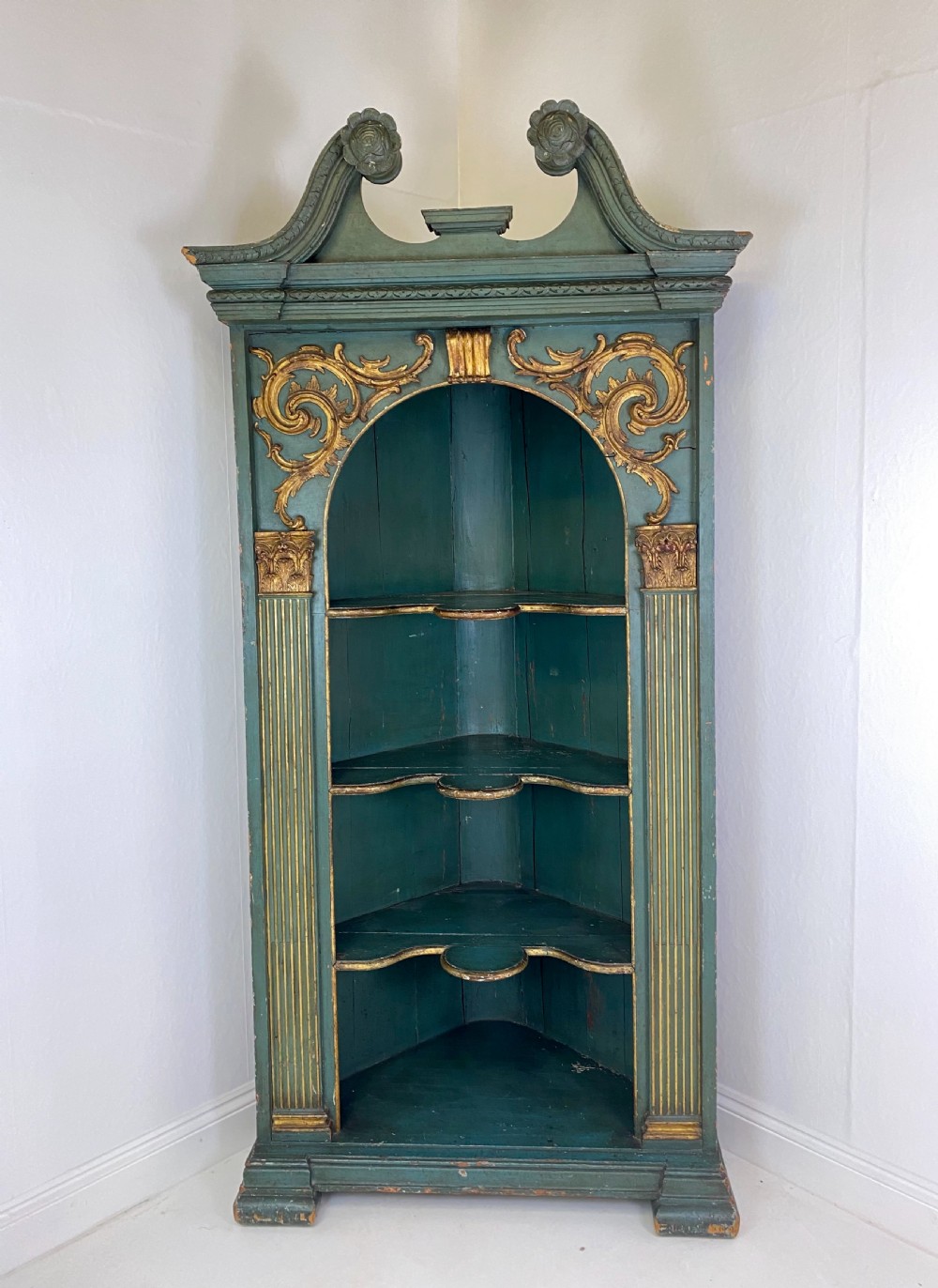 An 18th-century floor-standing teal green open corner cupboard featuring gold leaf detailing, fluted pilasters, and a broken swan-neck pediment.