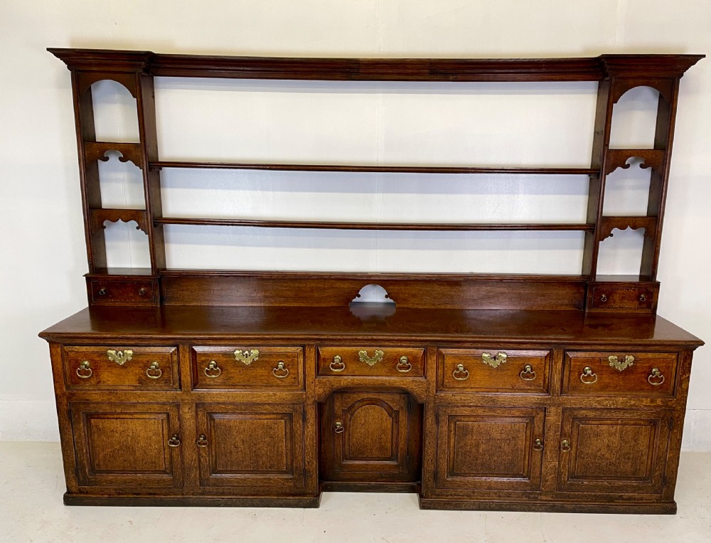 A large dark oak dresser featuring a multi-tiered open plate rack top above a wide base with drawers and panelled cupboard doors.