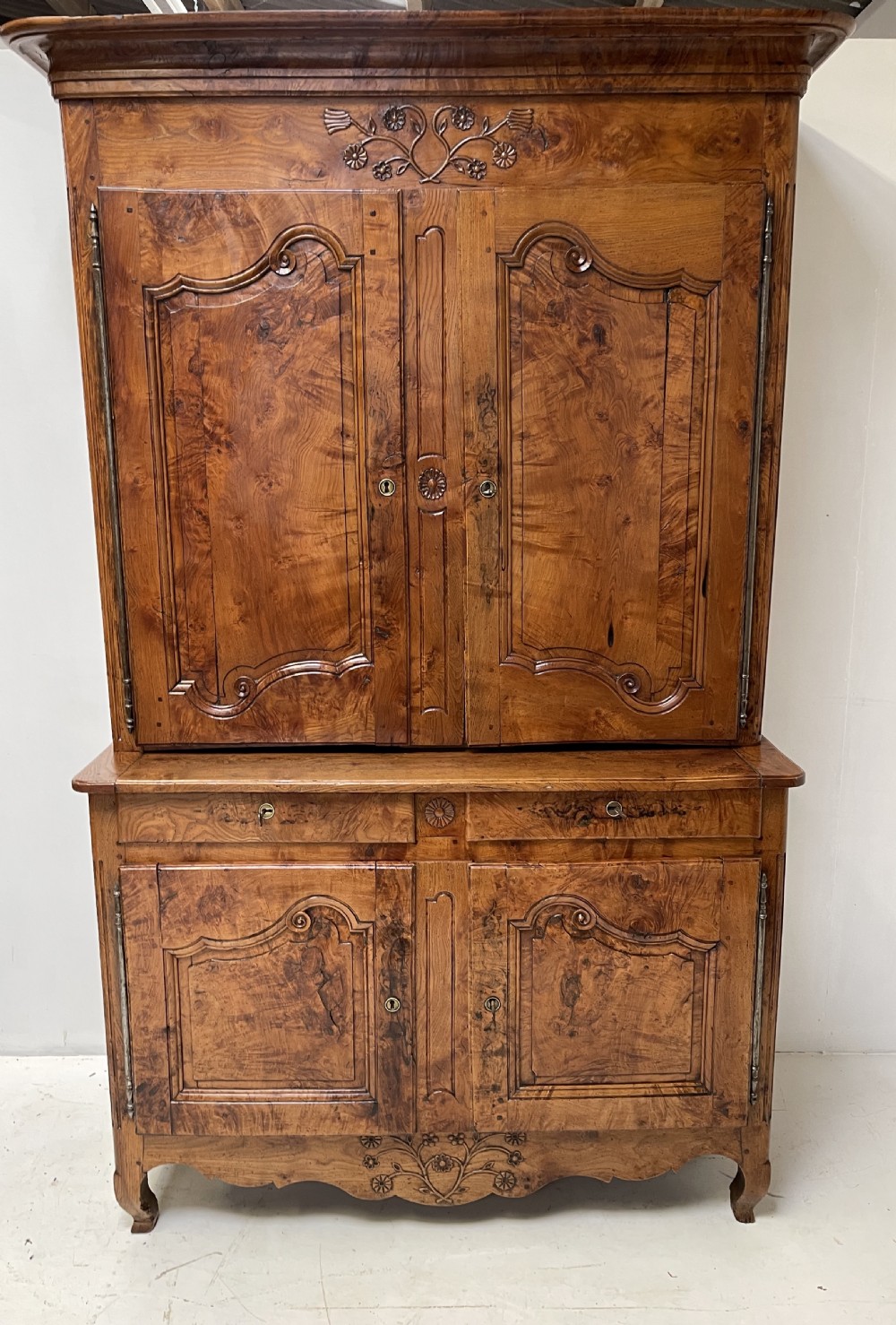 An antique two-piece French walnut cabinet featuring carved floral crests, molded panel doors, two drawers, and short cabriole feet.