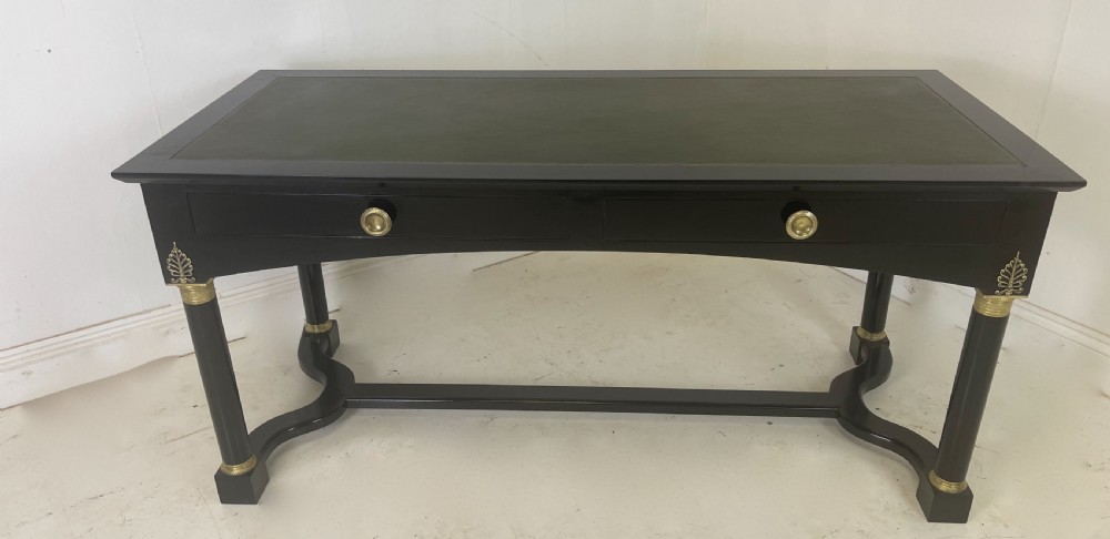 A black ebonised Empire style writing desk featuring a green leather inset top, two frieze drawers with brass knobs, ornate brass mounts, and cylindrical legs connected by a lower stretcher.