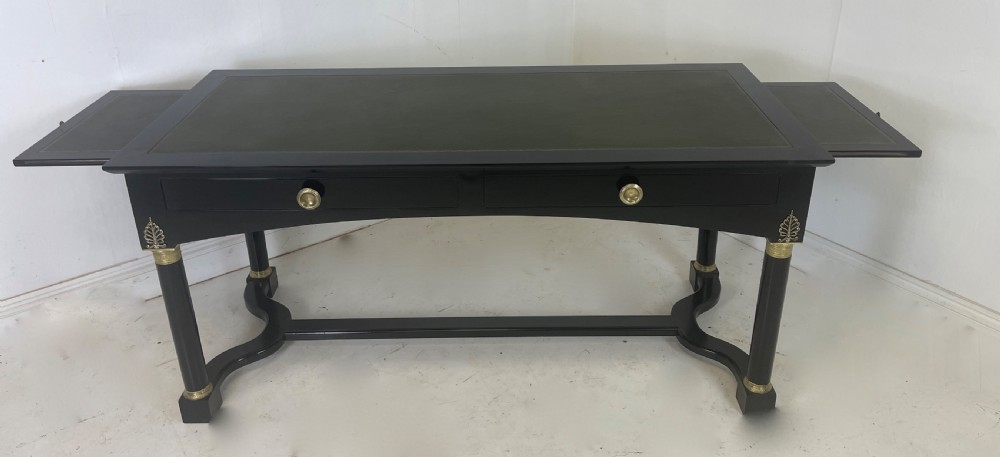 A black ebonised Empire style writing desk featuring a green leather inset top, two frieze drawers with brass knobs, ornate brass mounts, and cylindrical legs connected by a lower stretcher. View II