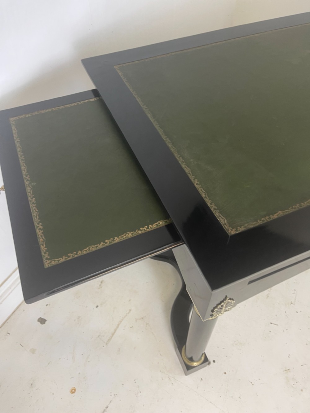 A black ebonised Empire style writing desk featuring a green leather inset top, two frieze drawers with brass knobs, ornate brass mounts, and cylindrical legs connected by a lower stretcher. View VII