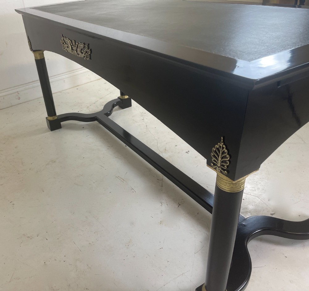 A black ebonised Empire style writing desk featuring a green leather inset top, two frieze drawers with brass knobs, ornate brass mounts, and cylindrical legs connected by a lower stretcher. View IX