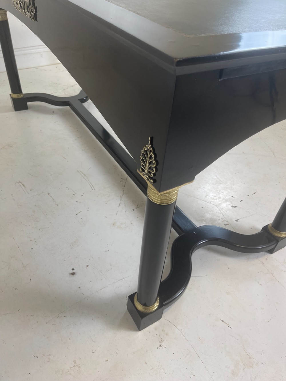 A black ebonised Empire style writing desk featuring a green leather inset top, two frieze drawers with brass knobs, ornate brass mounts, and cylindrical legs connected by a lower stretcher. View X