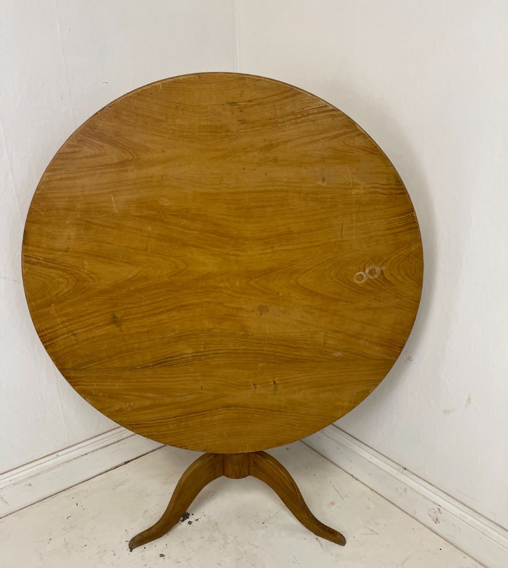 A circular pine tilt-top table presented in its raised vertical position showing its natural timber grain and tripod base.